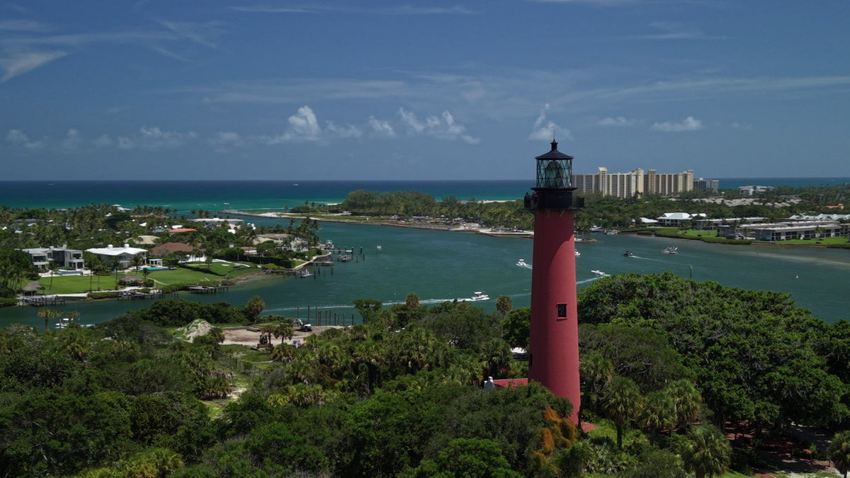 Aerial view of a red lighthouse surrounded by dense green trees, with turquoise water, boats, and coastal buildings under a blue sky. You will receive this 4K video which is 10sec. 